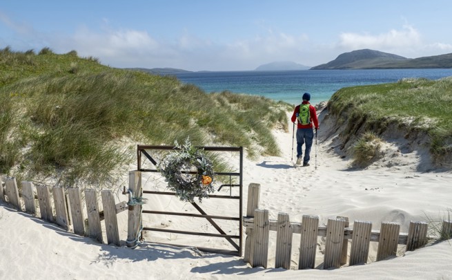 man walking on the sand with two sticks towards the sea with a gate closed behind him
