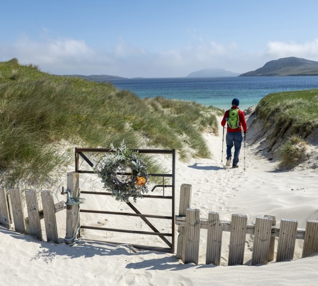 man walking on the sand with two sticks towards the sea with a gate closed behind him