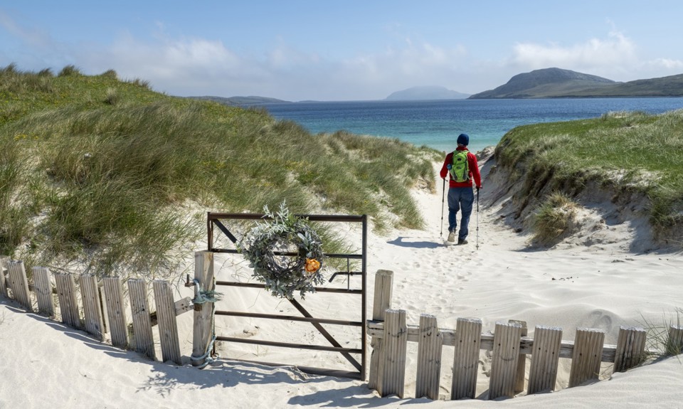 man walking on the sand with two sticks towards the sea with a gate closed behind him