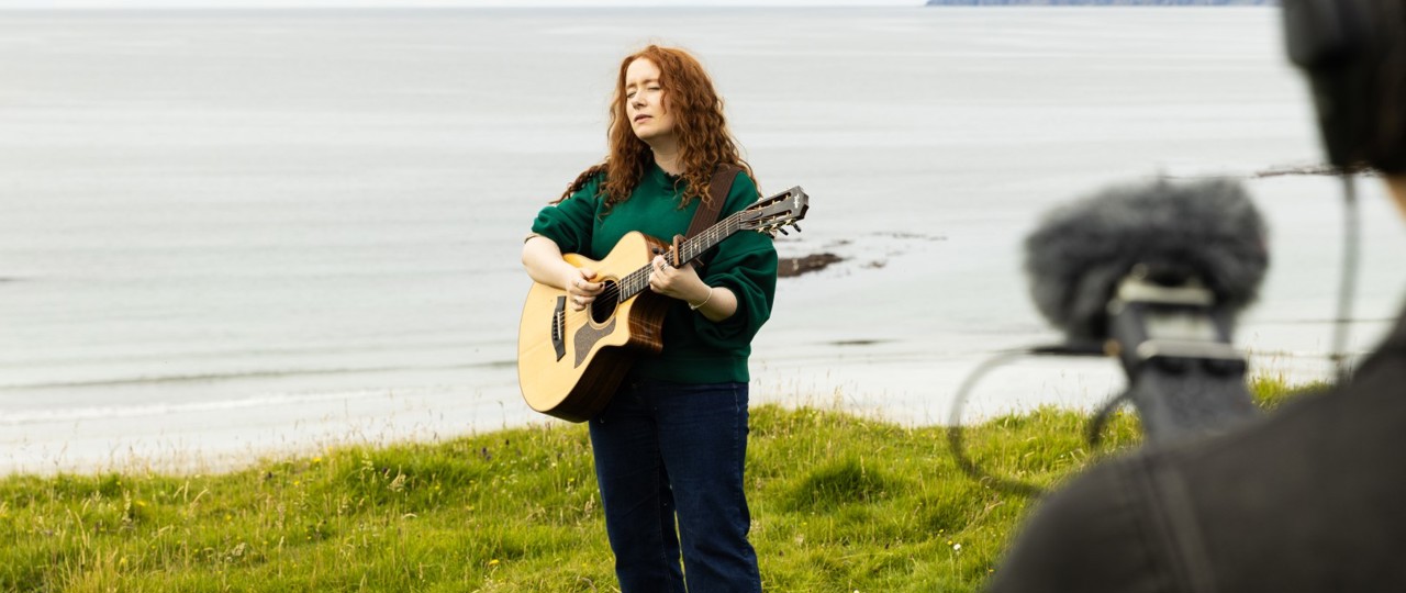 Nati Galloway singing on Eigg while playing a guitar the water is behind her