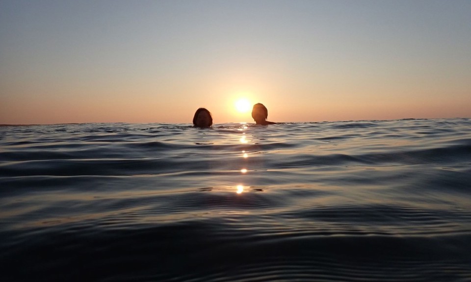 Two people are swimming in the ocean at sunset. The sun is low on the horizon, casting a warm glow across the sky and reflecting off the water. 