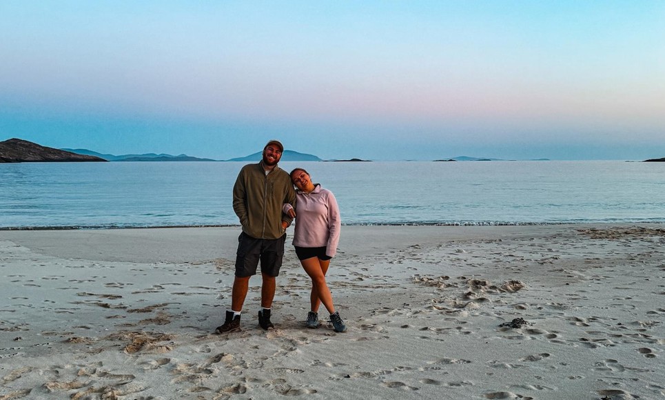 Two people stand on sandy beach during sunset or sunrise, with calm sea and distant islands in the background.