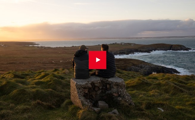 Two men sitting on a rock looking at the sunset in the distance, with a YouTube play button overlayed