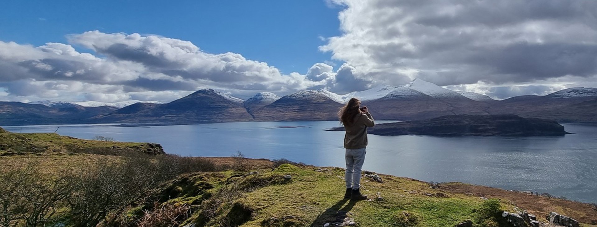 Person standing on a grassy hill overlooking a calm loch with snow-capped mountains under a partly cloudy sky.