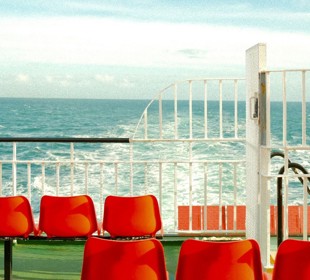 Red seats on outer deck of ferry with white railings and the sea in the background