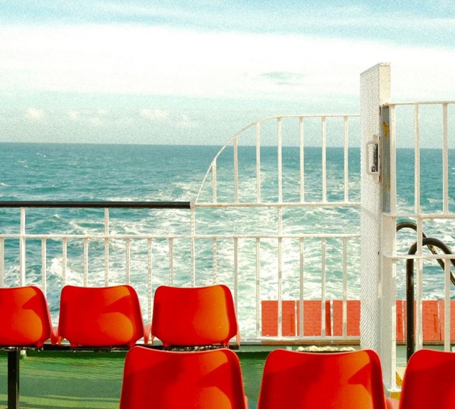 Red seats on outer deck of ferry with white railings and the sea in the background