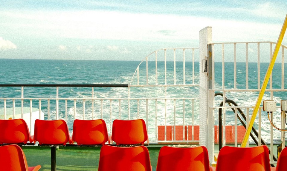 Red seats on outer deck of ferry with white railings and the sea in the background