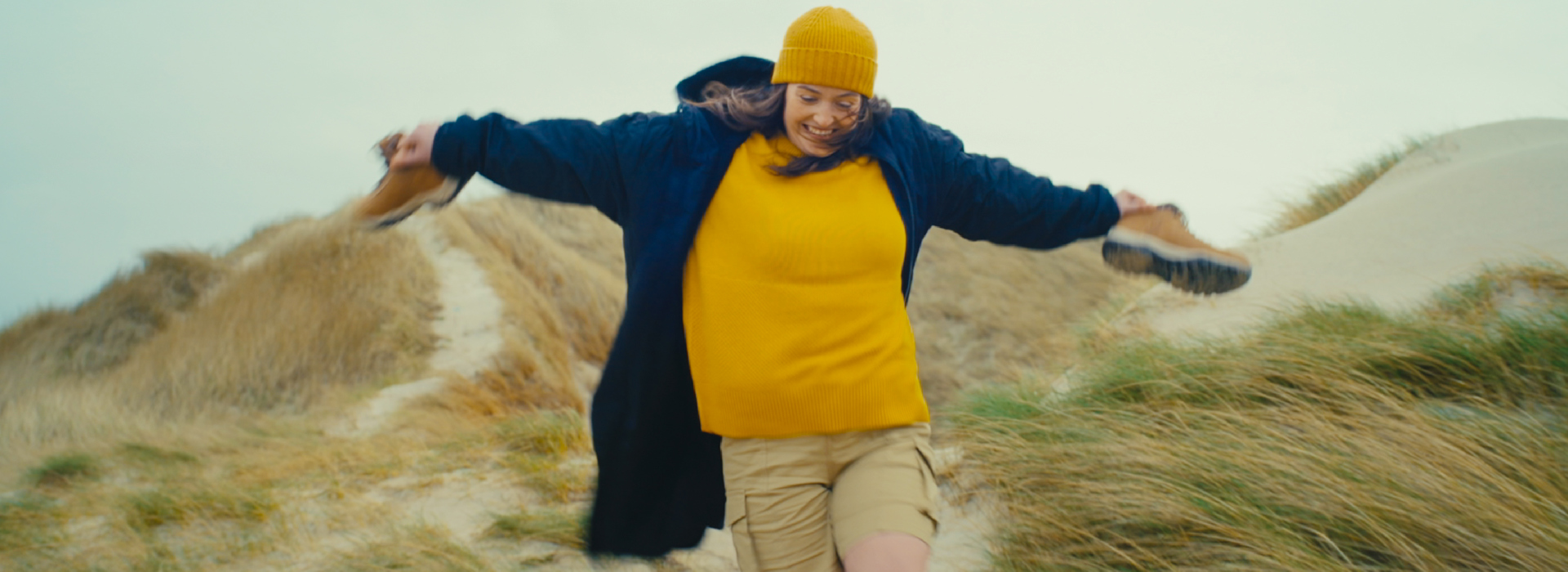 Brunette female with yellow hat with boots in her hand, running barefoot down a grassy and sandy sand dune