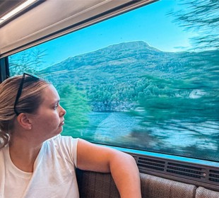 A woman with blonde hair looks out train window at scenic landscape with mountains and trees.
