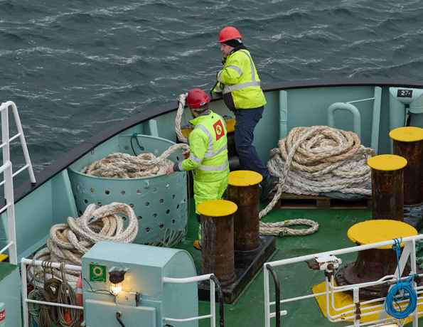 Two members of the CalMac crew, in high visibiity clothing, working at the bow of the ship with rope.