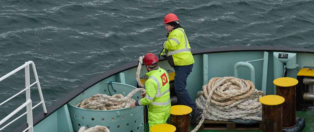 Two members of the CalMac crew, in high visibiity clothing, working at the bow of the ship with rope.