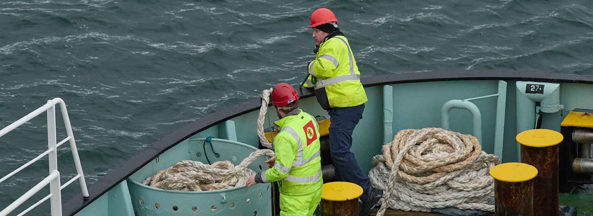 Two members of the CalMac crew, in high visibiity clothing, working at the bow of the ship with rope.