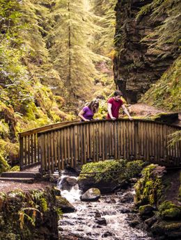 A couple looking over a small bridge as water runs underneath at the wooded area called Pucks Glen