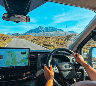 Person driving a Ford on a scenic road with mountains ahead, using GPS navigation on the dashboard screen.