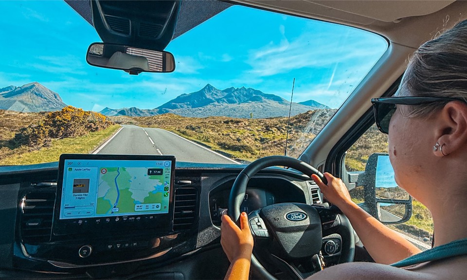 Person driving a Ford on a scenic road with mountains ahead, using GPS navigation on the dashboard screen.