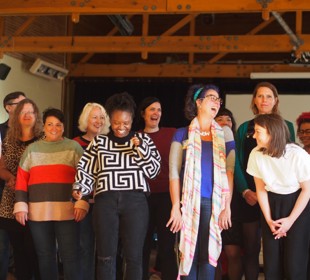 A group of people standing in a room with wooden ceiling beams. A projector screen and lighting equipment are visible in the background.