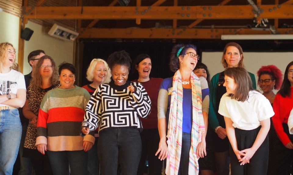 A group of people standing in a room with wooden ceiling beams. A projector screen and lighting equipment are visible in the background.