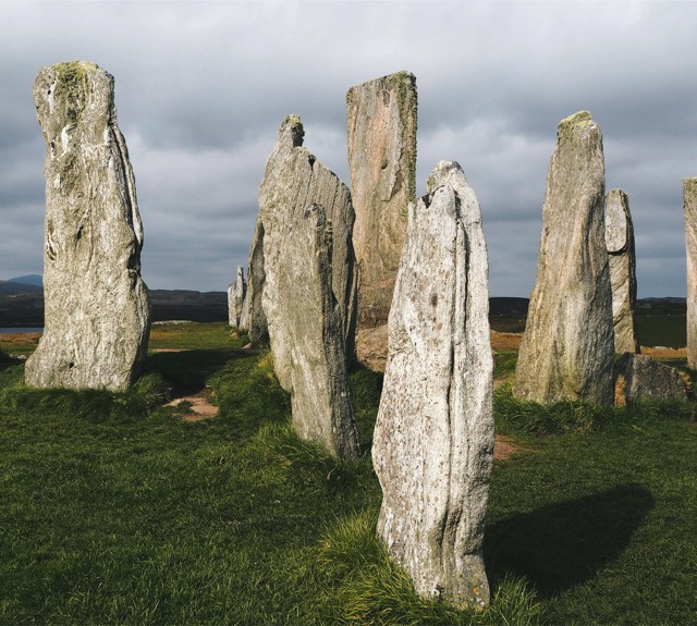 Circle of tall, ancient standing stones on a grassy field beneath a cloudy sky.