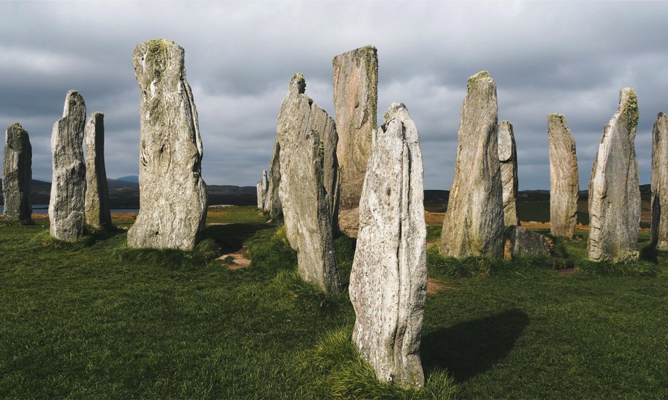 Circle of tall, ancient standing stones on a grassy field beneath a cloudy sky.