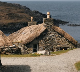 Stone cottages with thatched roofs by the sea, surrounded by rocky terrain and a gravel path.