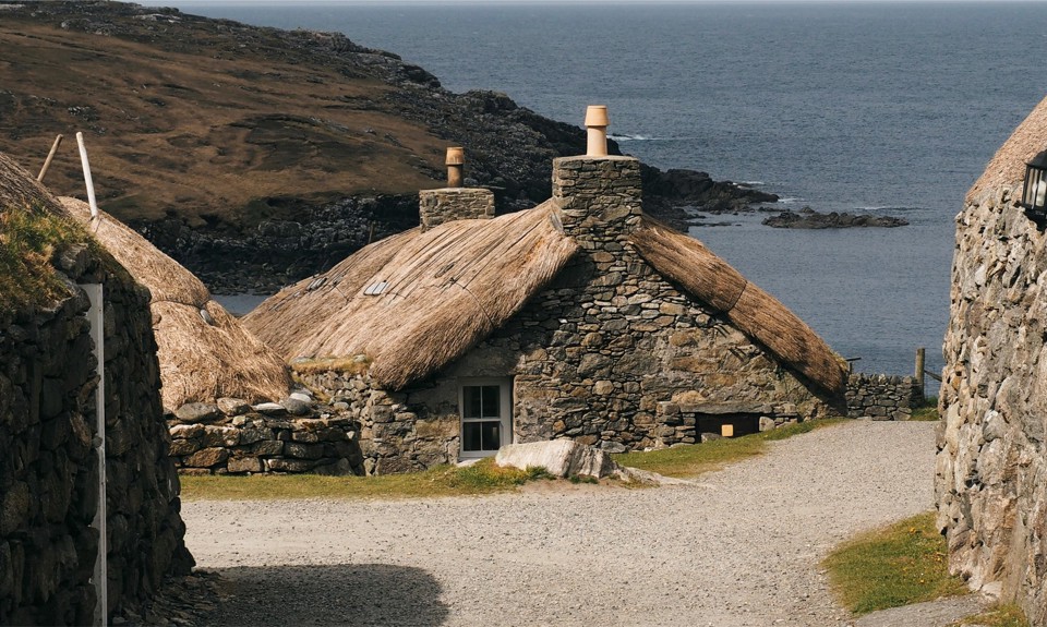 Stone cottages with thatched roofs by the sea, surrounded by rocky terrain and a gravel path.