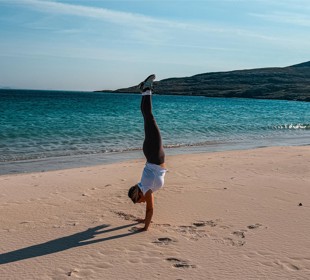 Person doing a handstand on a sandy beach with ocean waves and hills in the background.