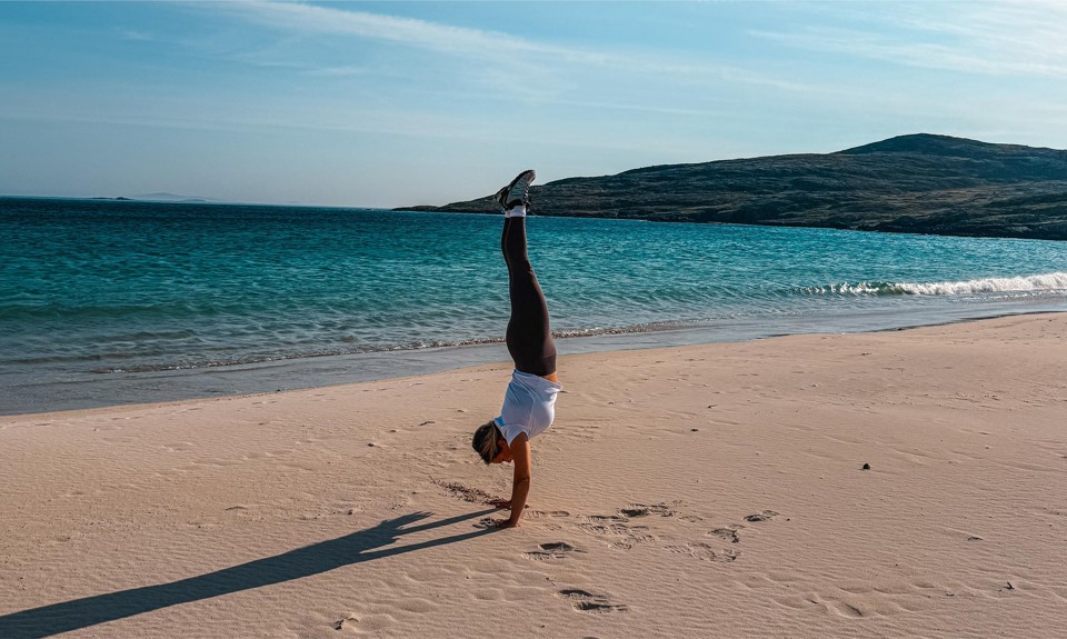 Person doing a handstand on a sandy beach with ocean waves and hills in the background.
