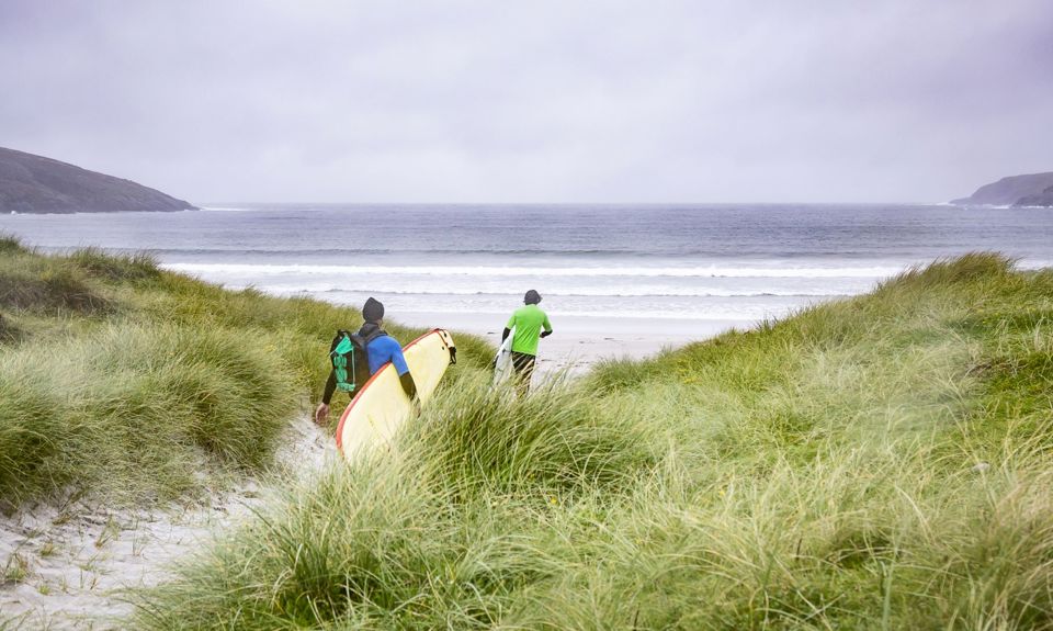 Surfers carrying a surfboard as they walk to a sandy beach.