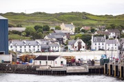 The village of Port Ellen with hillside houses, greenery, and a dock area featuring trucks and stacked logs in the foreground.