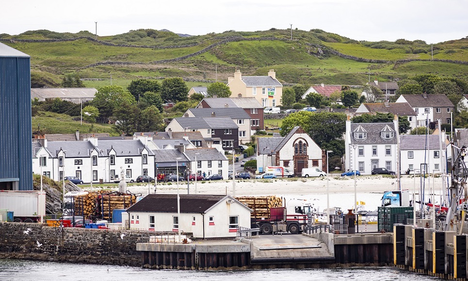 The village of Port Ellen with hillside houses, greenery, and a dock area featuring trucks and stacked logs in the foreground.