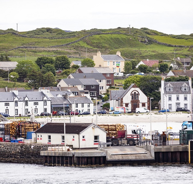 The village of Port Ellen with hillside houses, greenery, and a dock area featuring trucks and stacked logs in the foreground.