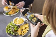 A person taking a photo of a meal with a mobile. The meal is fries, macaroni and cheese, with salad, the plate in the background has fish and chips.