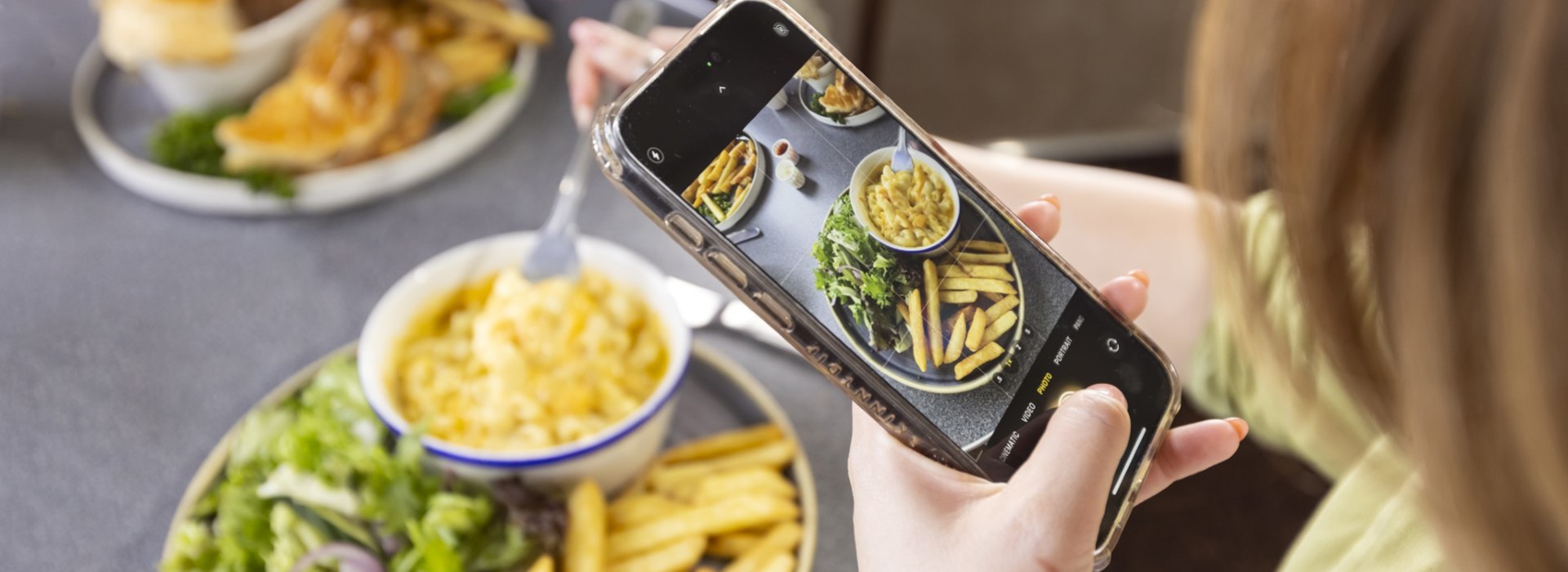 A person taking a photo of a meal with a mobile. The meal is fries, macaroni and cheese, with salad, the plate in the background has fish and chips.