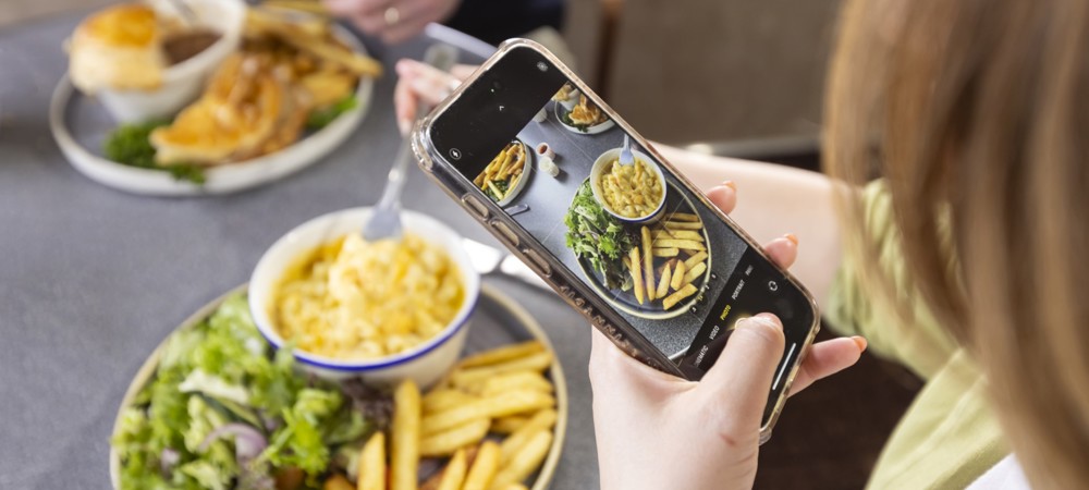A person taking a photo of a meal with a mobile. The meal is fries, macaroni and cheese, with salad, the plate in the background has fish and chips.