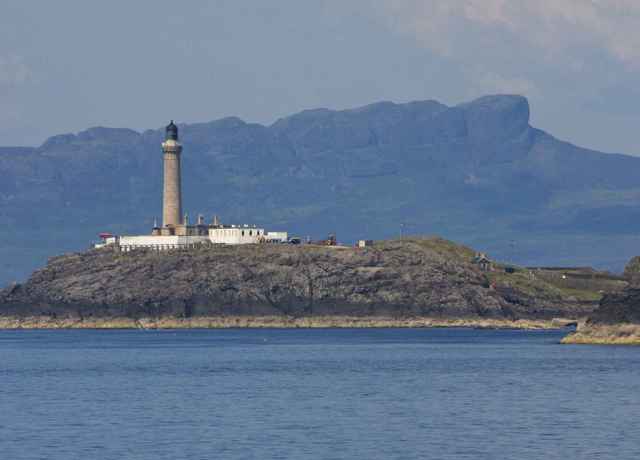 Ardnamurn Lighthouse sitting on a rocky peak above the water with mountainous shape of the Sgurr of Eigg in background.