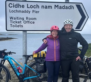 Two cyclists arm-in-arm at Lochmaddy Pier sign, marking a stop on their Hebridean cycling journey.