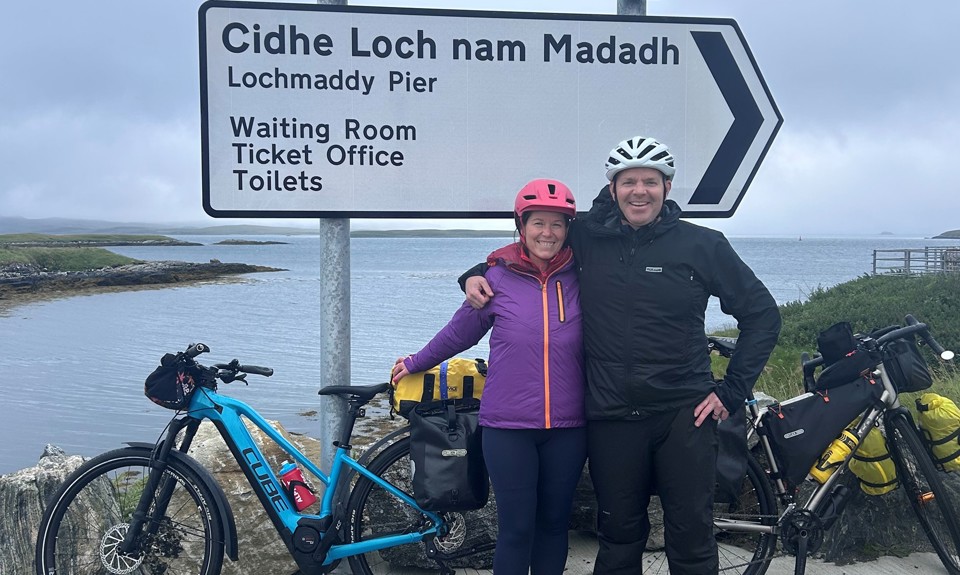 Two cyclists arm-in-arm at Lochmaddy Pier sign, marking a stop on their Hebridean cycling journey.