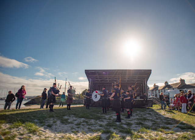 A pipe band performing in front of a small stage at a local festival