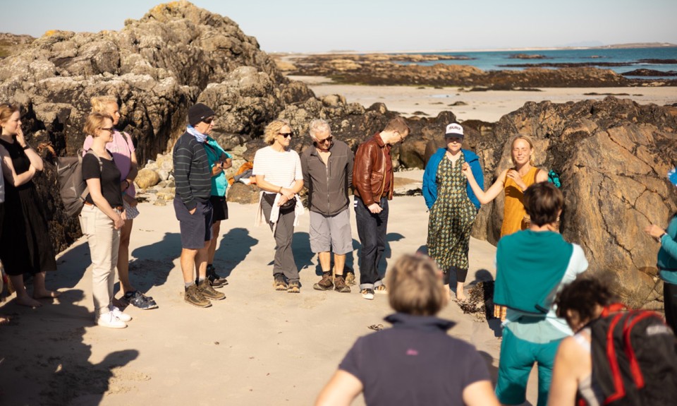 A group of people standing on a rocky beach. The background features large rock formations and the ocean with a clear sky.