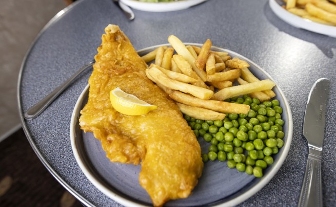 A plate with a large piece of battered fish topped with a lemon wedge, fries, and peas. 