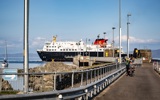 Caledonian MacBrayne ferry docked at a pier, with people walking and cycling nearby and a 'PEDESTRIANS' sign visible on a sunny day.