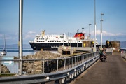 Caledonian MacBrayne ferry docked at a pier, with people walking and cycling nearby and a 'PEDESTRIANS' sign visible on a sunny day.