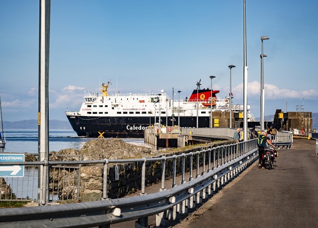 Caledonian MacBrayne ferry docked at a pier, with people walking and cycling nearby and a 'PEDESTRIANS' sign visible on a sunny day.
