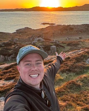 Eddie taking a selfie on a rocky coastal hillside at sunset, overlooking calm sea waters and distant islands.