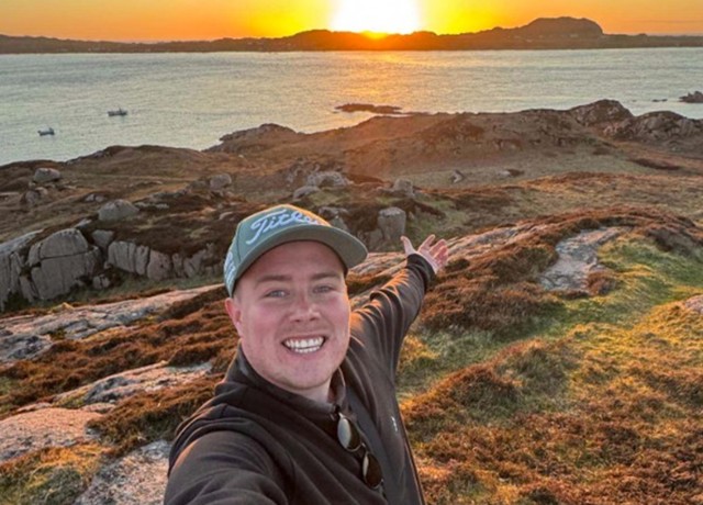 Eddie taking a selfie on a rocky coastal hillside at sunset, overlooking calm sea waters and distant islands.