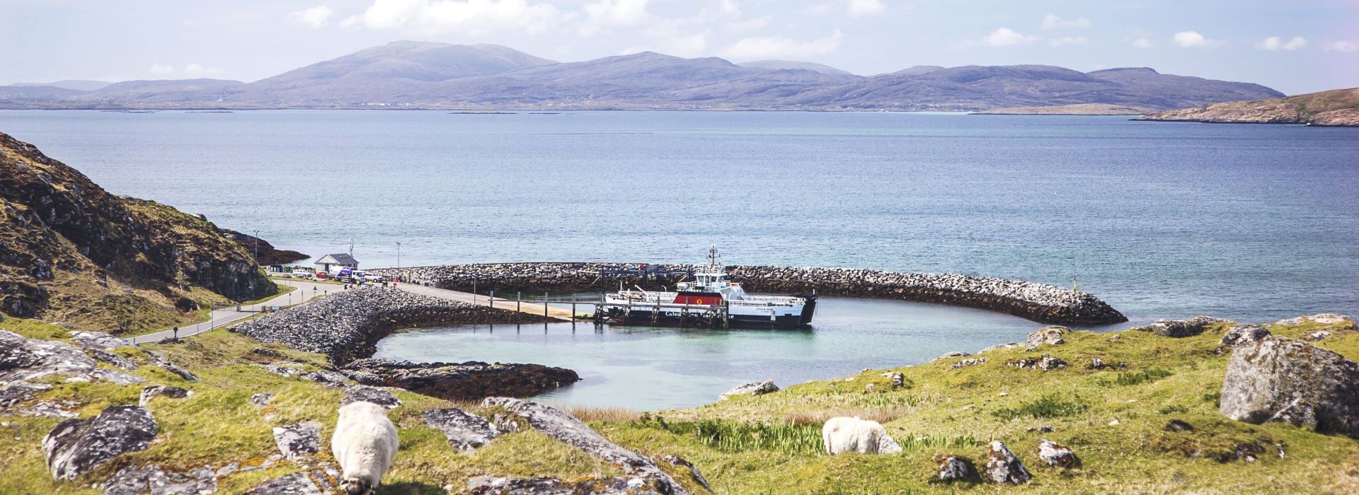 A view from a hilltop of a CalMac ferry arriving at Eriskay, with rolling hills across a body of water in the background.