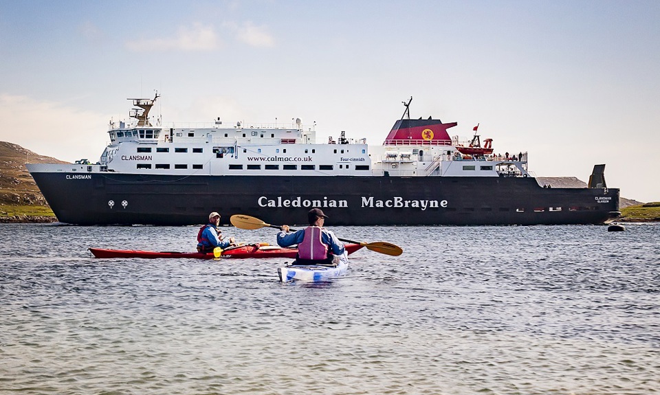 Two people rowing in kayaks with the CalMac ferry MV Clansman in sailing past in the background.