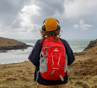 Brunette female with yellow hat, black over ear headphones, red backpack looking out to the sea. 