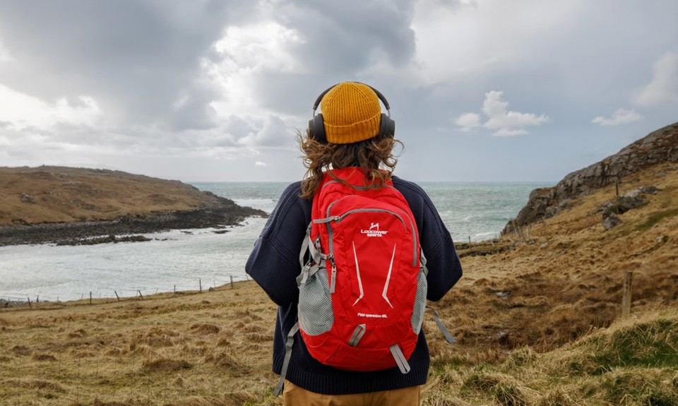 Brunette female with yellow hat, black over ear headphones, red backpack looking out to the sea. 