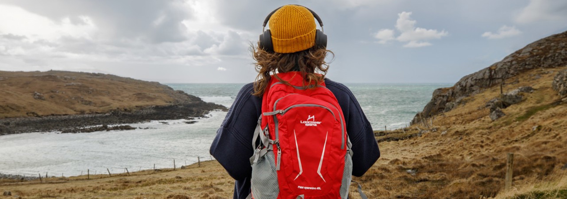 Brunette female with yellow hat, black over ear headphones, red backpack looking out to the sea. 